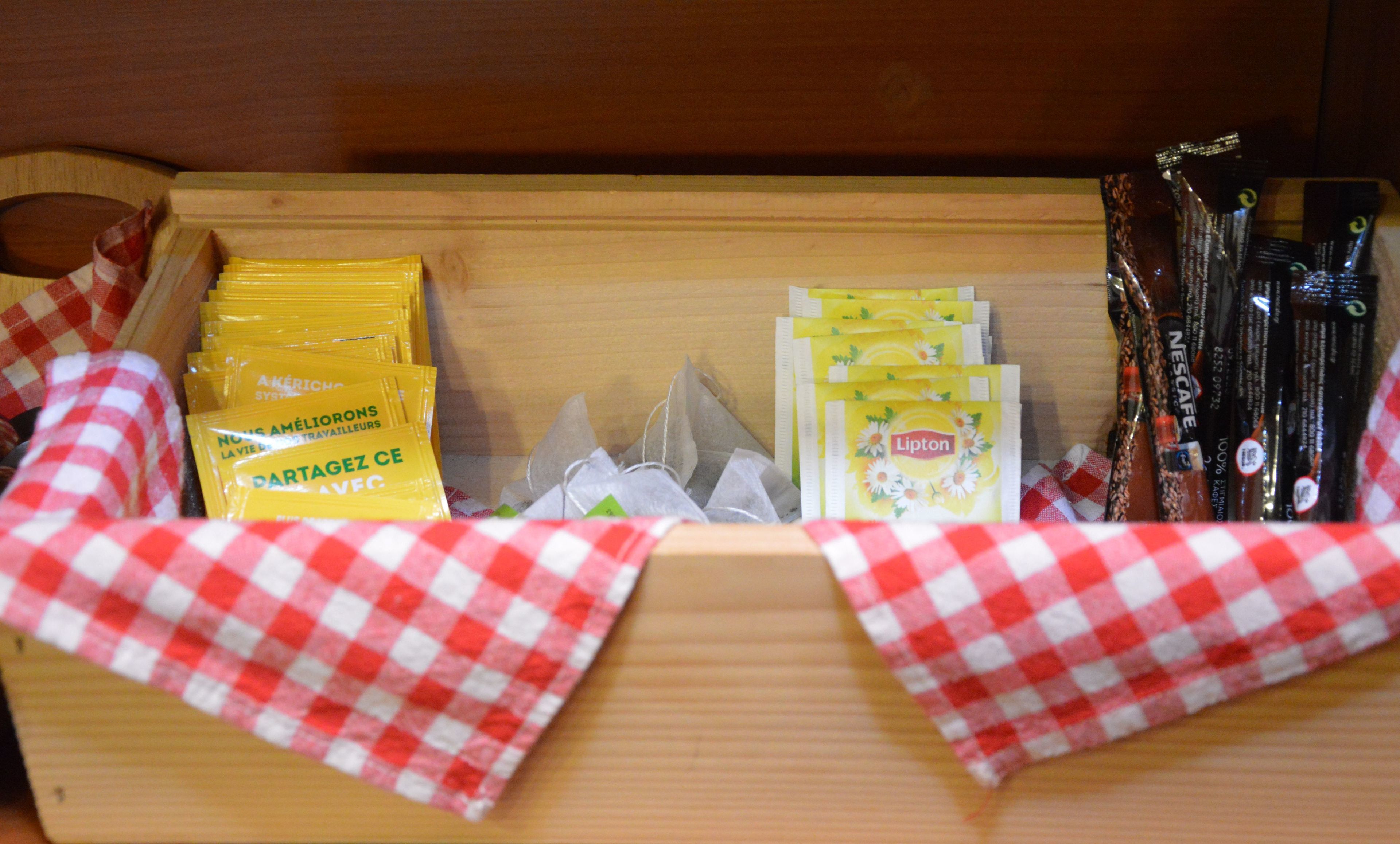 Puinen laatikko, jossa on erilaisia elintarvikkeita, kuten Liptonin teepusseja ja Nescafén kahvipaketteja, aseteltuna punavalkoiseen ruutukankaaseen.