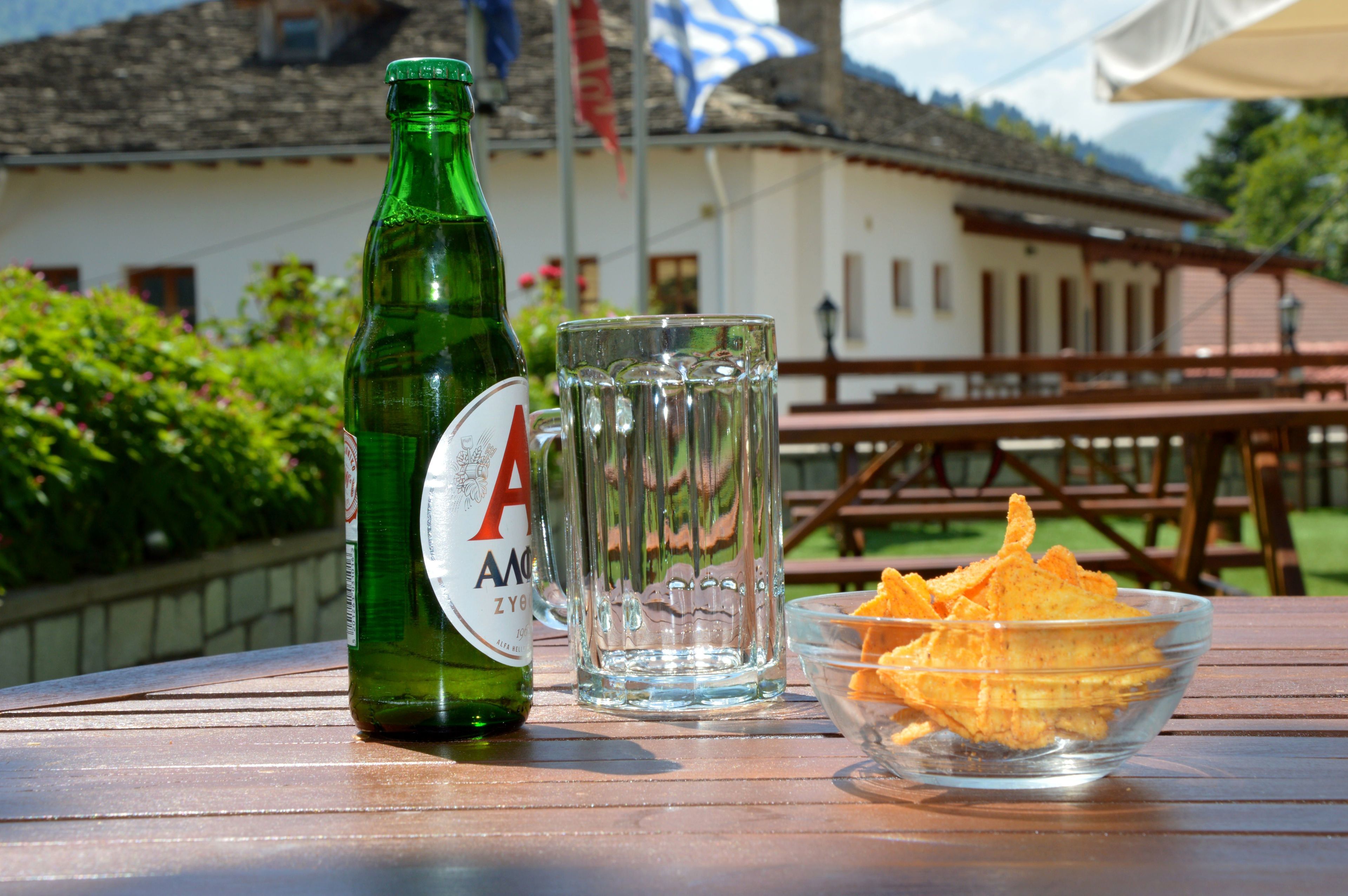 A green bottle, a glass, and a bowl of chips on a wooden table.