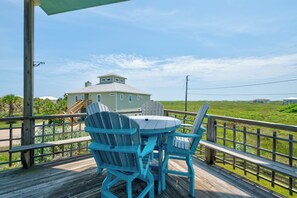 Um deck de madeira com duas cadeiras Adirondack e uma mesa, com vista para uma paisagem gramada e uma casa com telhado em gable.