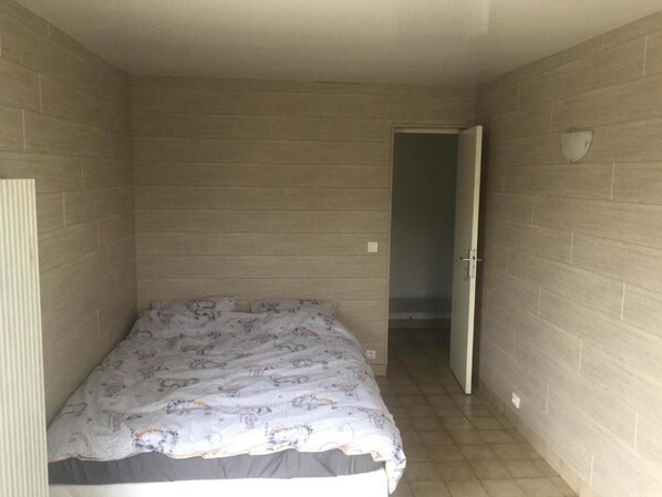 Una habitación pequeña y estrecha con una cama apoyada en la pared izquierda, una puerta que da a otra habitación y una lámpara en la pared derecha.