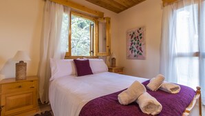 A neatly made bed with a purple blanket and two rolled towels, a wooden nightstand with a lamp, and a window with curtains. - Kyveli Cottage (Foini)