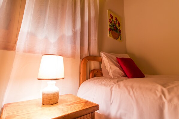 Una cama bien tendida con almohadas blancas y rojas, una mesita de noche de madera con una lámpara, y un cuadro de flores en la pared.