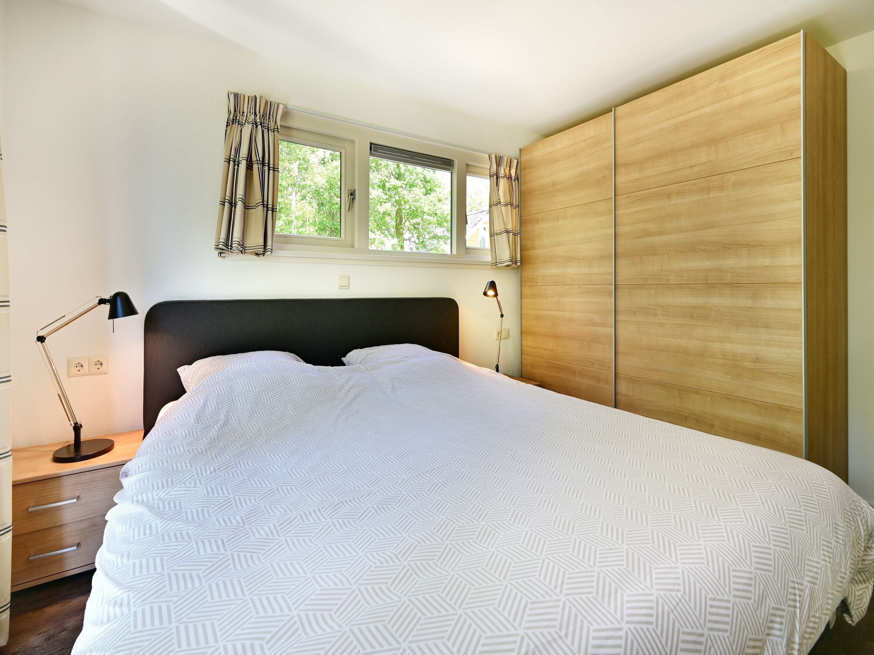 Un dormitorio con cama, mesita de noche, lámpara, ventana con cortinas y un armario de madera.