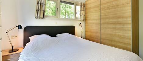 Un dormitorio con cama, mesita de noche, lámpara, ventana con cortinas y un armario de madera.