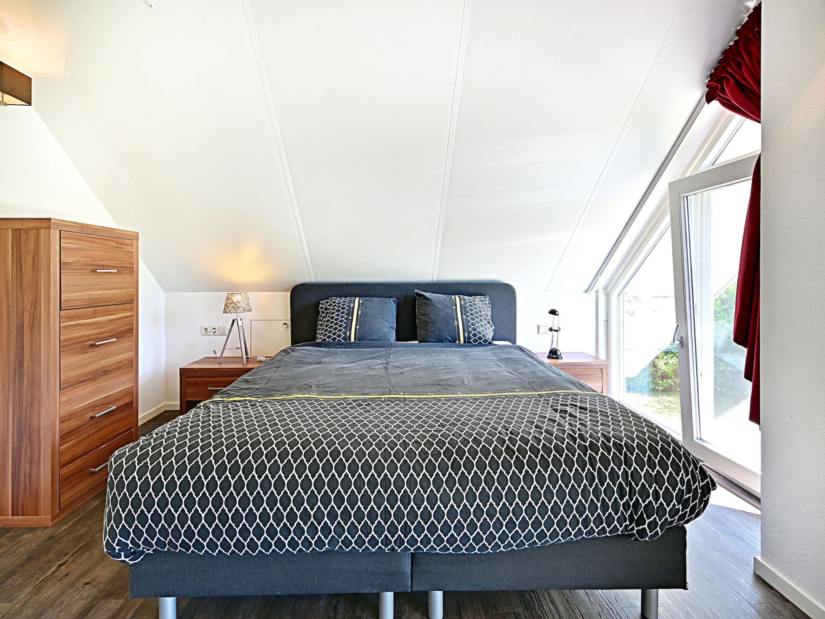 A bedroom with a bed, a wooden dresser, a nightstand, a lamp, and a window with curtains.