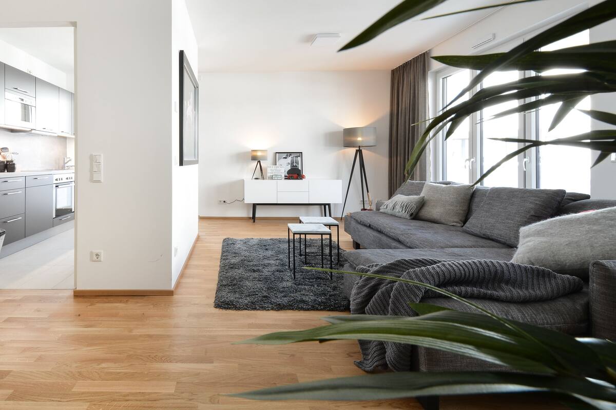 Sala de estar moderna com sofá cinza, uma mesinha lateral e uma luminária de chão. Cozinha visível ao fundo, com armários brancos e micro-ondas.