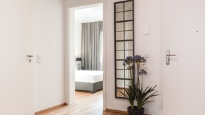 A hallway with a wooden floor leading to a bedroom with a bed and curtains. There is a potted plant with purple flowers and a tall, narrow mirror on the wall. - Lunas Appartements in Essen (Essen)