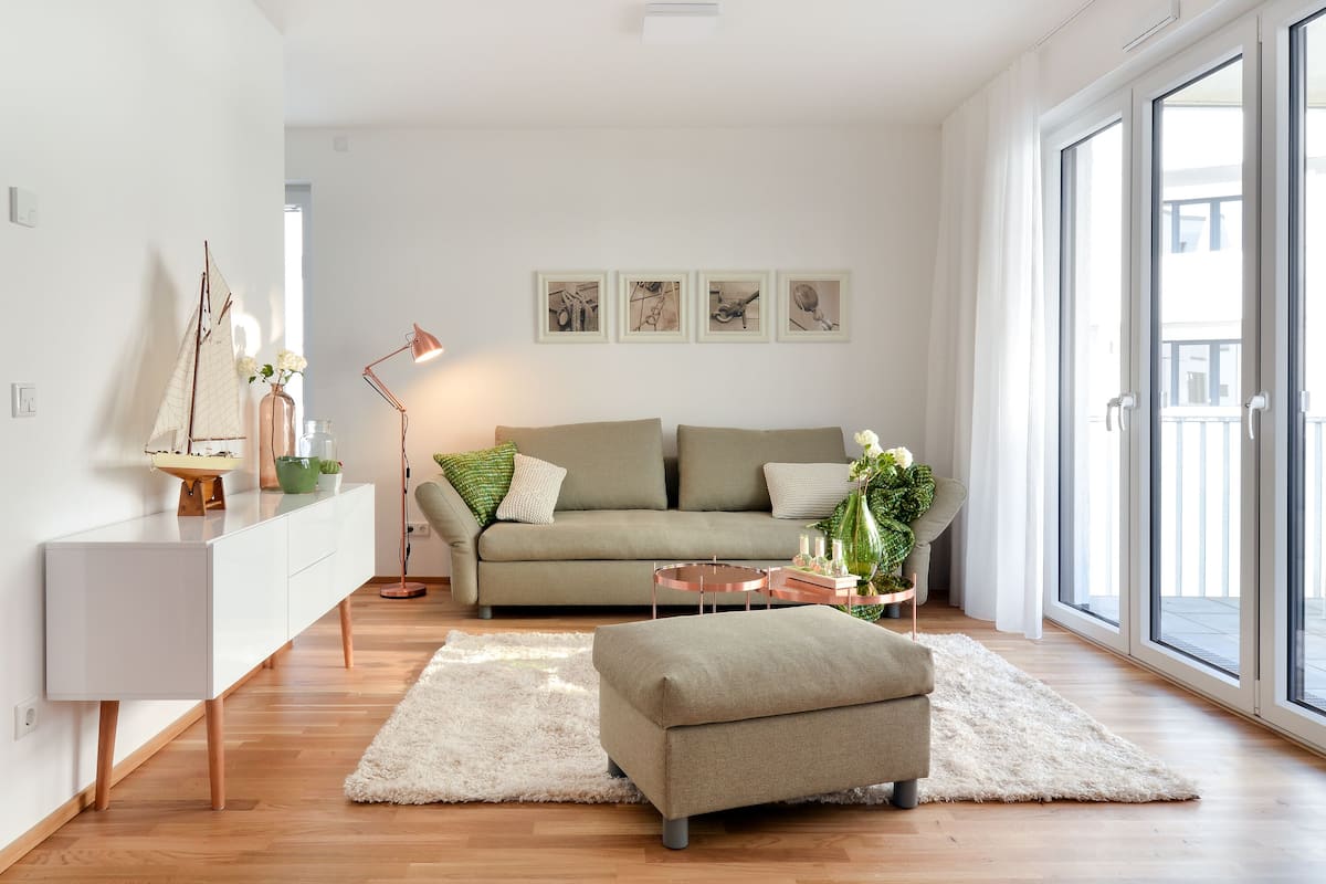 Uma sala de estar moderna com um sofá, um banquinho e uma mesa de centro. Há um armário branco, um abajur e uma obra de arte na parede.