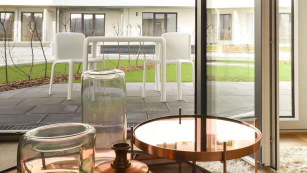 A modern room with a glass door leading to an outdoor seating area with white chairs and a glass table. - Lunas Appartements in Essen (Essen)