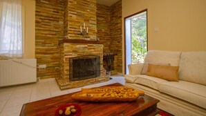 Una sala de estar con chimenea de piedra, una mesa de centro de madera con frutos secos, un sofá blanco y una ventana con vistas a la vegetación.