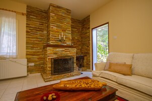 Una sala de estar con chimenea de piedra, una mesa de centro de madera con frutos secos, un sofá blanco y una ventana con vistas a la vegetación.