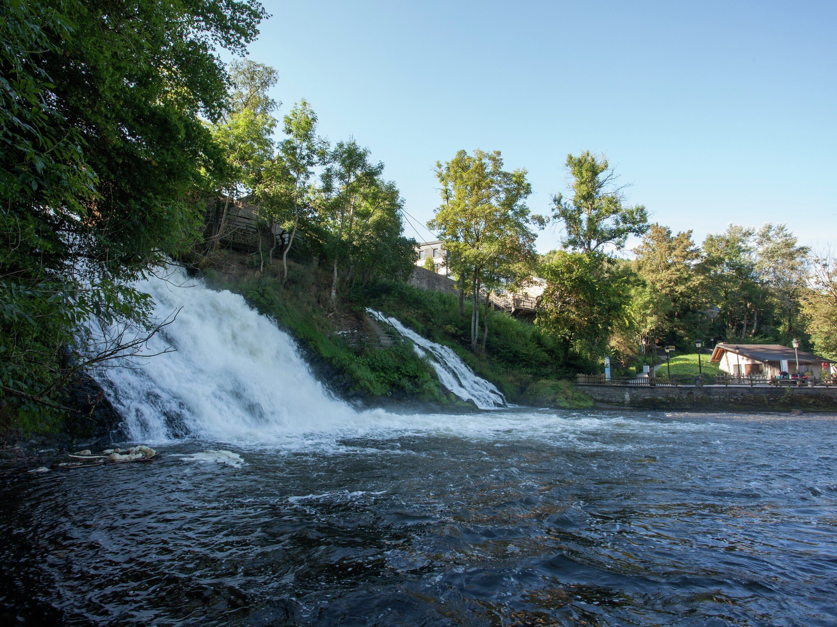 A waterfall with a river in the foreground, surrounded by greenery and a small building in the background.