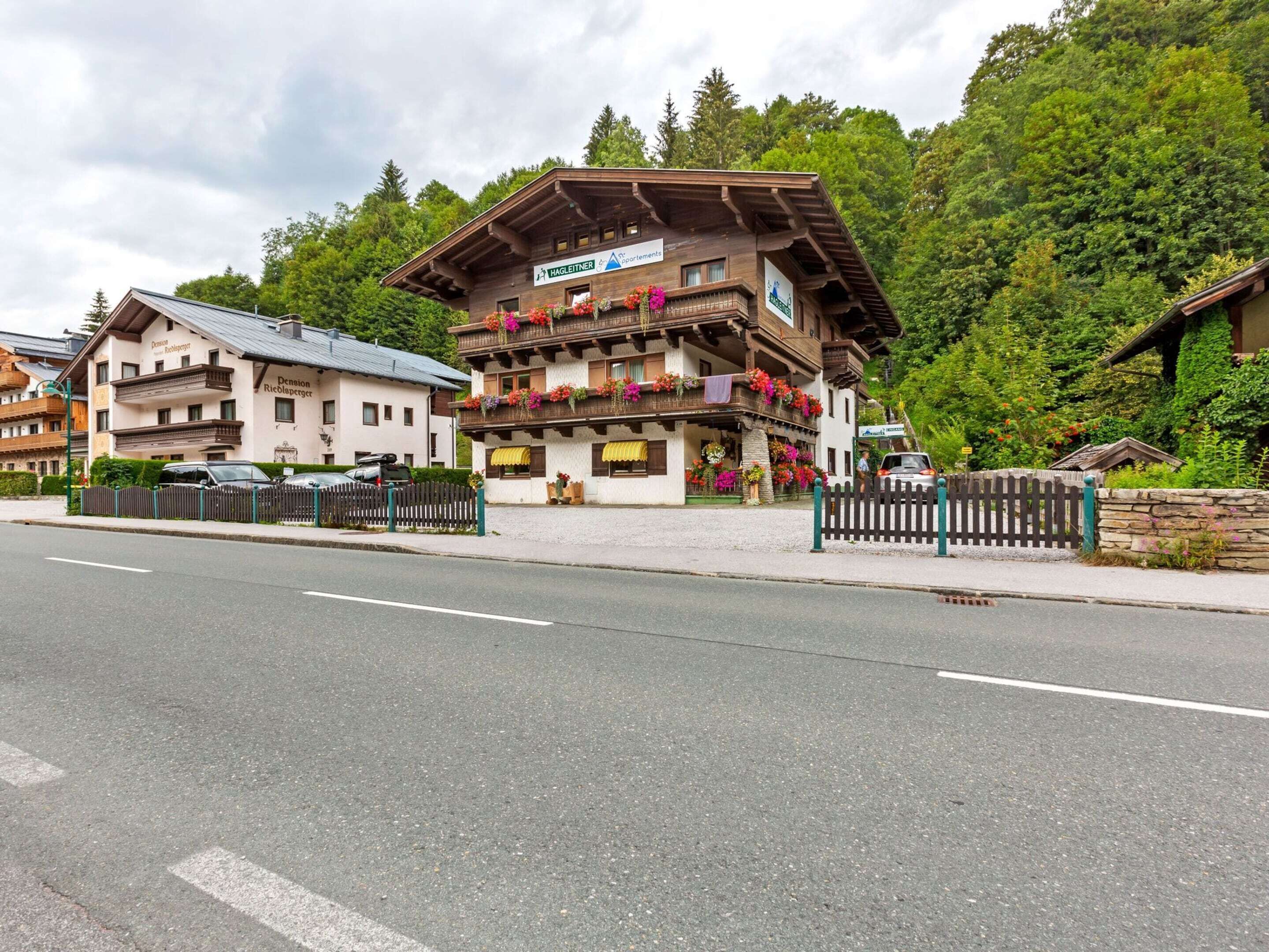 A traditional wooden building with balconies adorned with flowers, a paved road in front, and a forested backdrop.