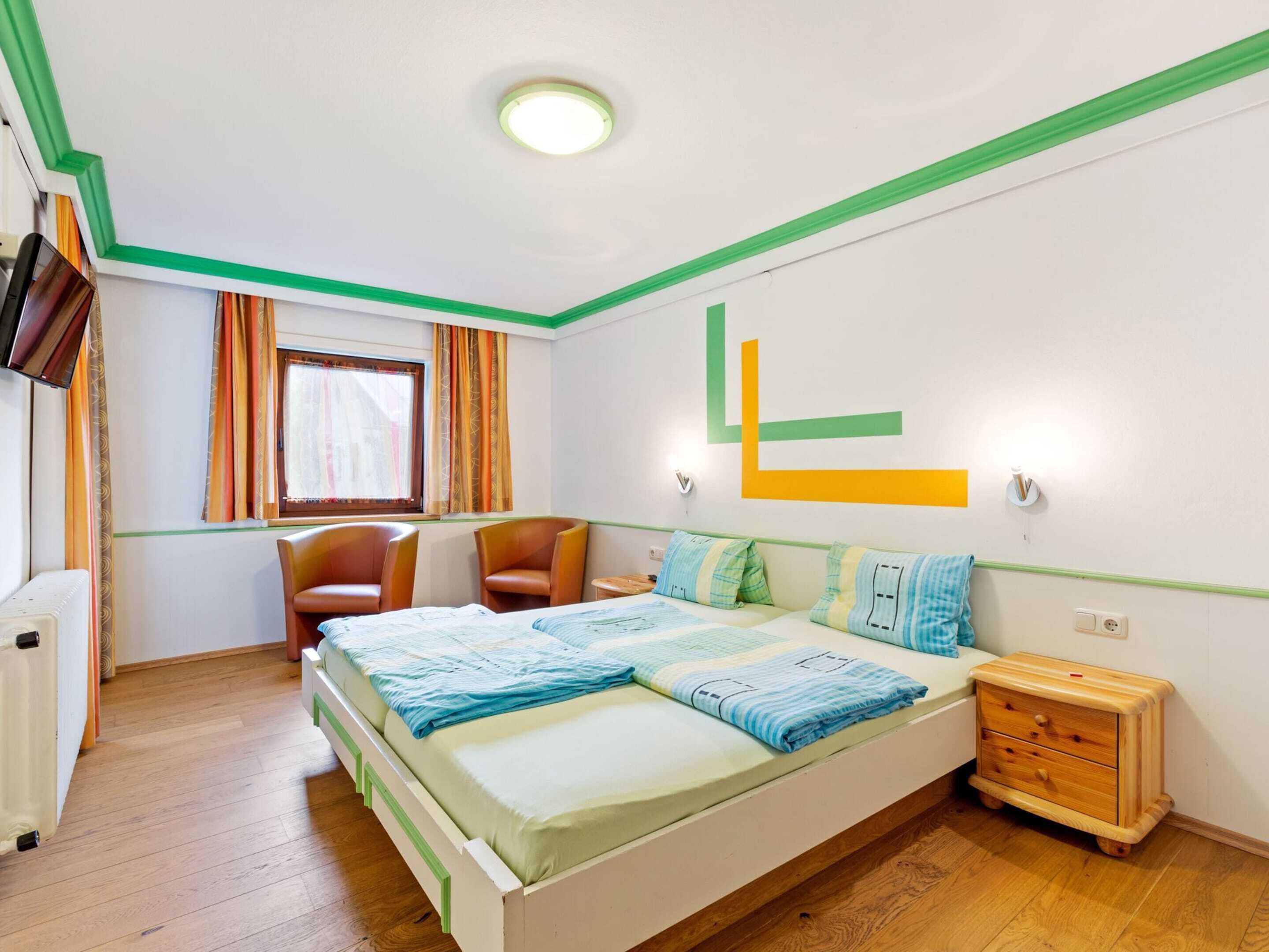 A bedroom with a bed, two chairs, a small wooden nightstand, a radiator, and a window with curtains.