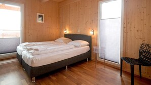 A bedroom with a wooden ceiling, a bed with white bedding, two wall-mounted lights, a chair, and a window with blinds.