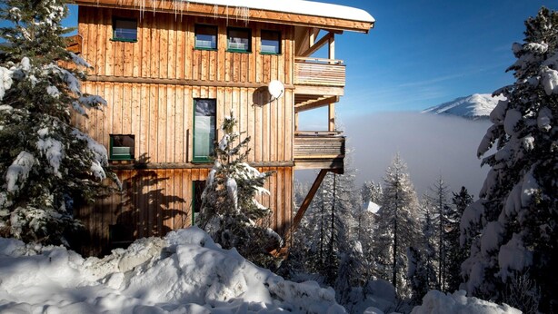 A wooden cabin with multiple windows and a balcony, surrounded by snow-covered trees and a mountain in the background.