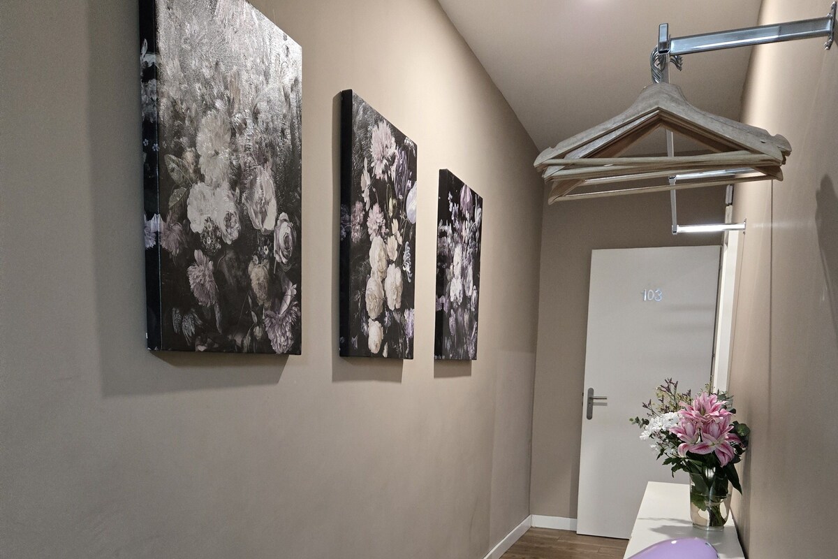 A hallway with three wall-mounted paintings, a door with a number, and a hanging coat rack.