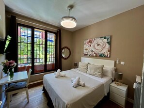 Habitación de hotel con cama, mesitas de noche, escritorio y un amplio ventanal con vistas a edificios y zonas verdes.