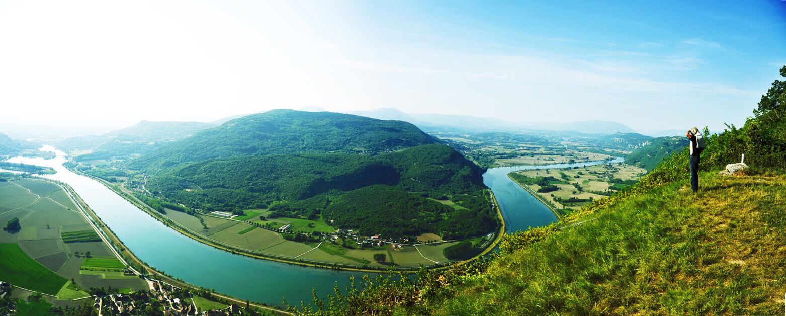 Vista panorâmica de um rio serpenteando por um vale com colinas, um pequeno povoado e uma pessoa fotografando.