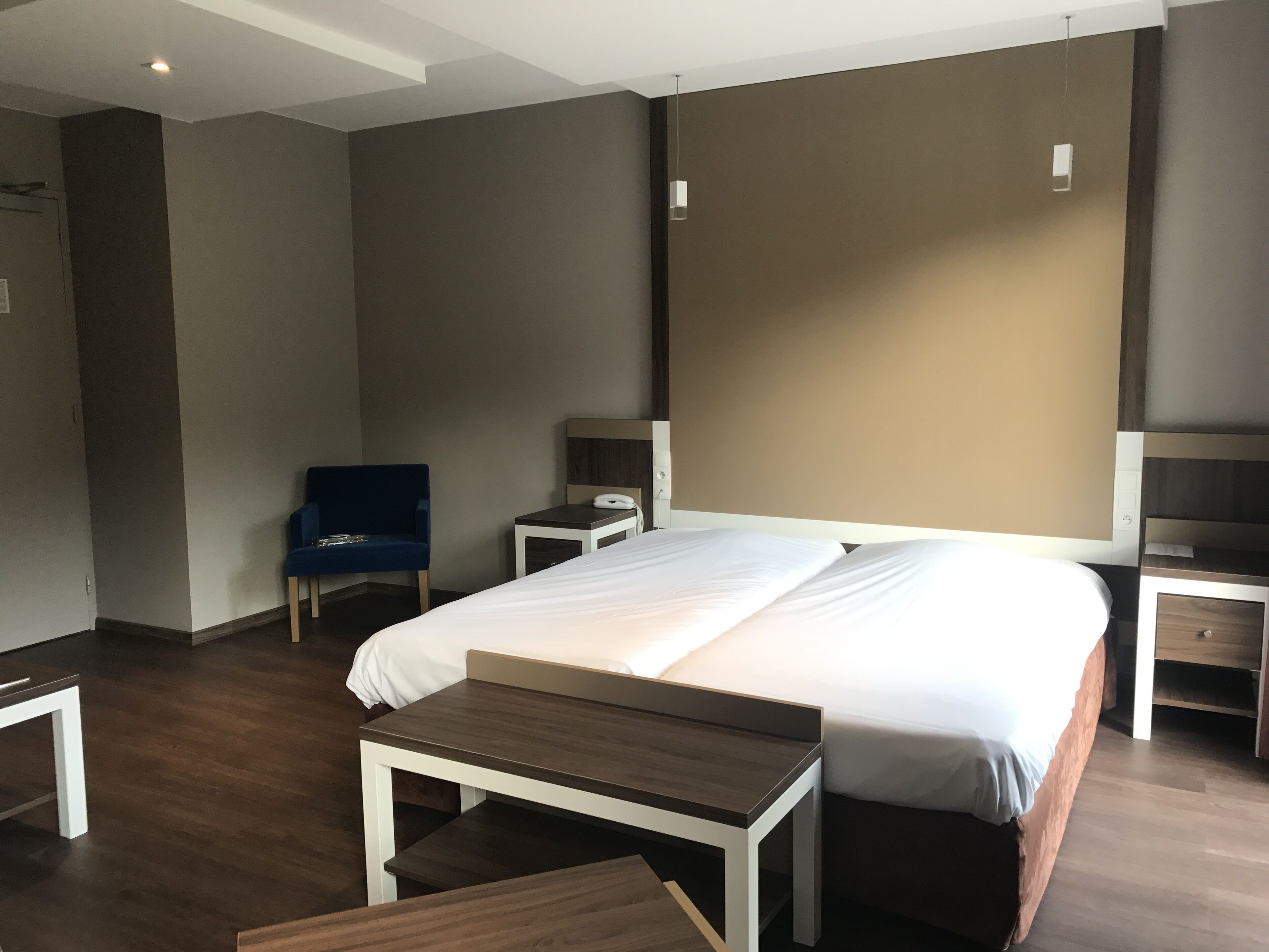 A hotel room with a large bed, a small desk, a chair, and a bedside table.