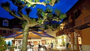 A courtyard with outdoor seating, a large tree, and a building with a balcony. - Hotel Restaurant Les Bergeronnettes (Champagneux)