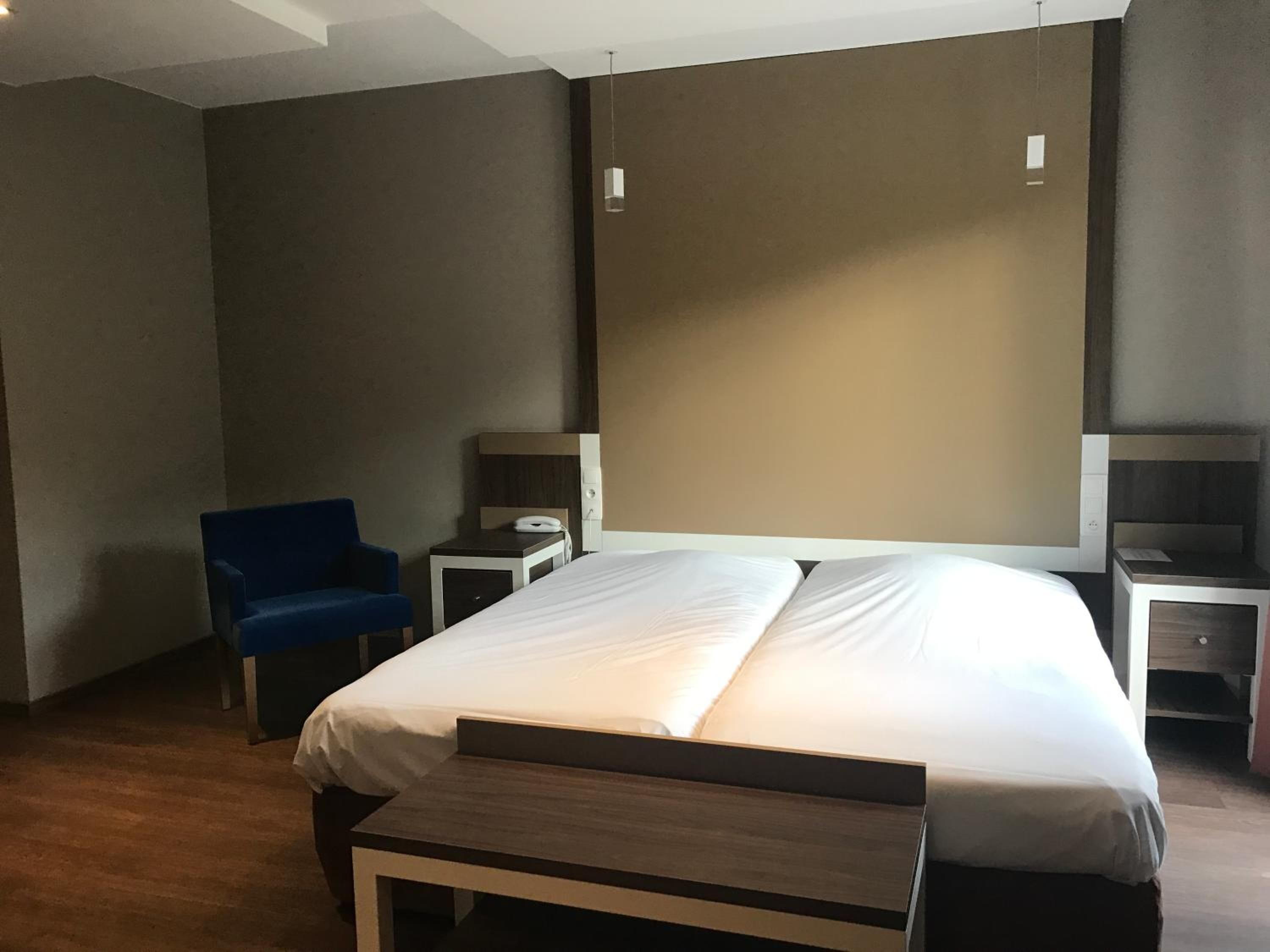 A hotel room with a large bed, a blue chair, a bedside table with a lamp, and a small desk with a chair.