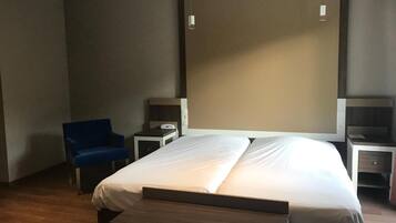 A hotel room with a large bed, a blue chair, a bedside table with a lamp, and a small desk with a chair.