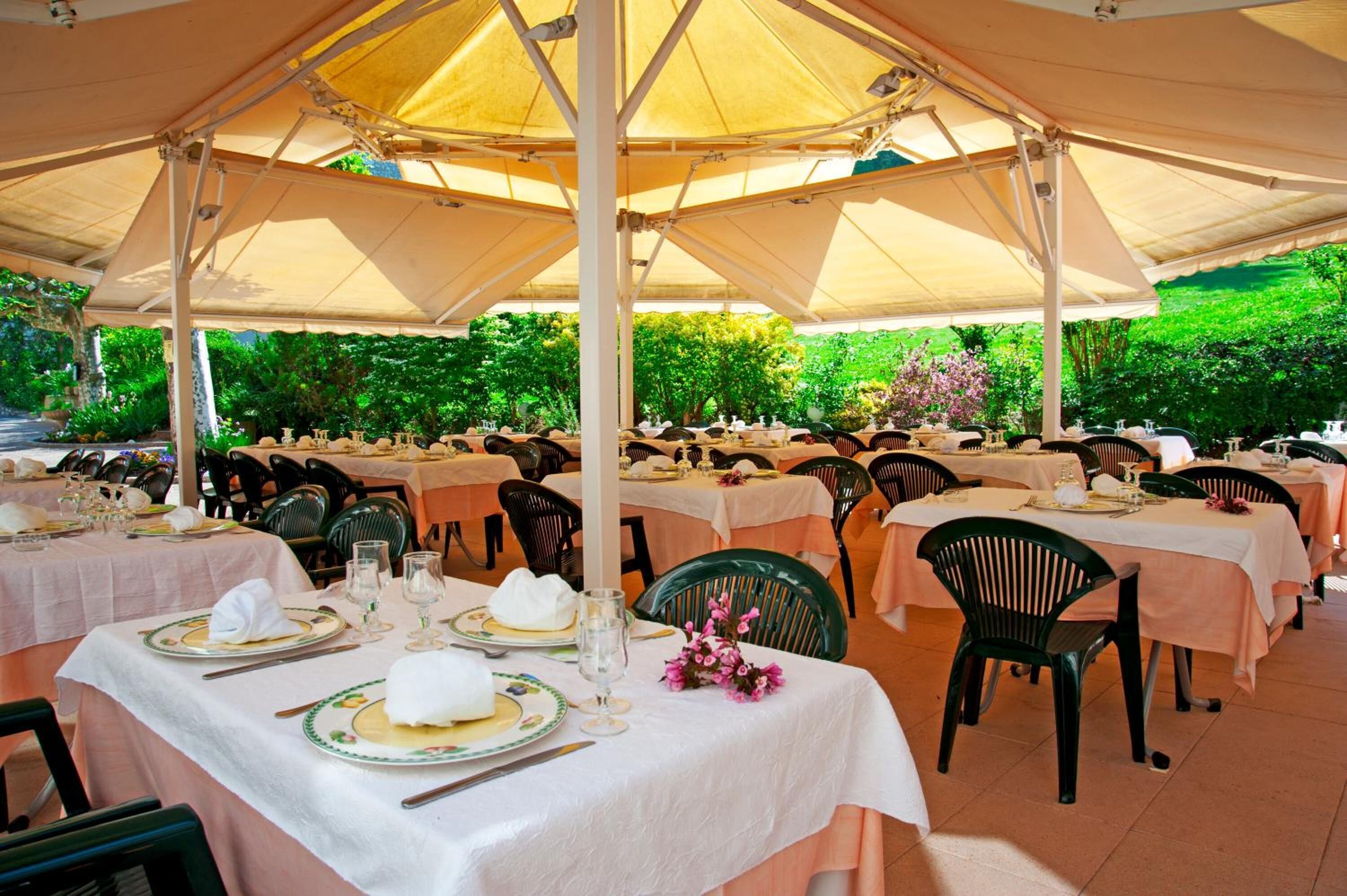 A covered outdoor dining area with tables set for a meal, surrounded by greenery.
