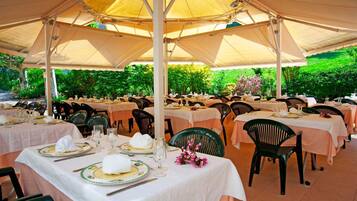 A covered outdoor dining area with tables set for a meal, surrounded by greenery.