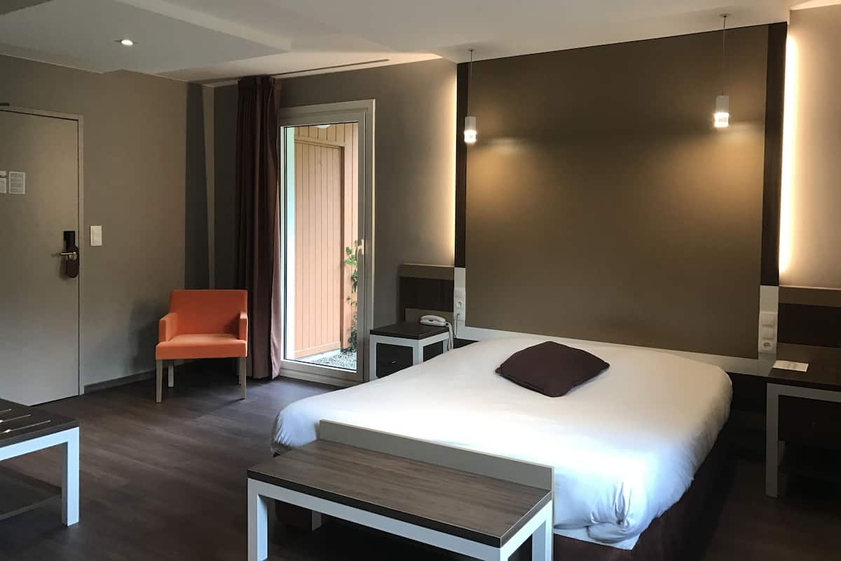 A hotel room with a bed, a desk, a chair, a bedside table, a nightstand, a telephone, and a door with a curtain.