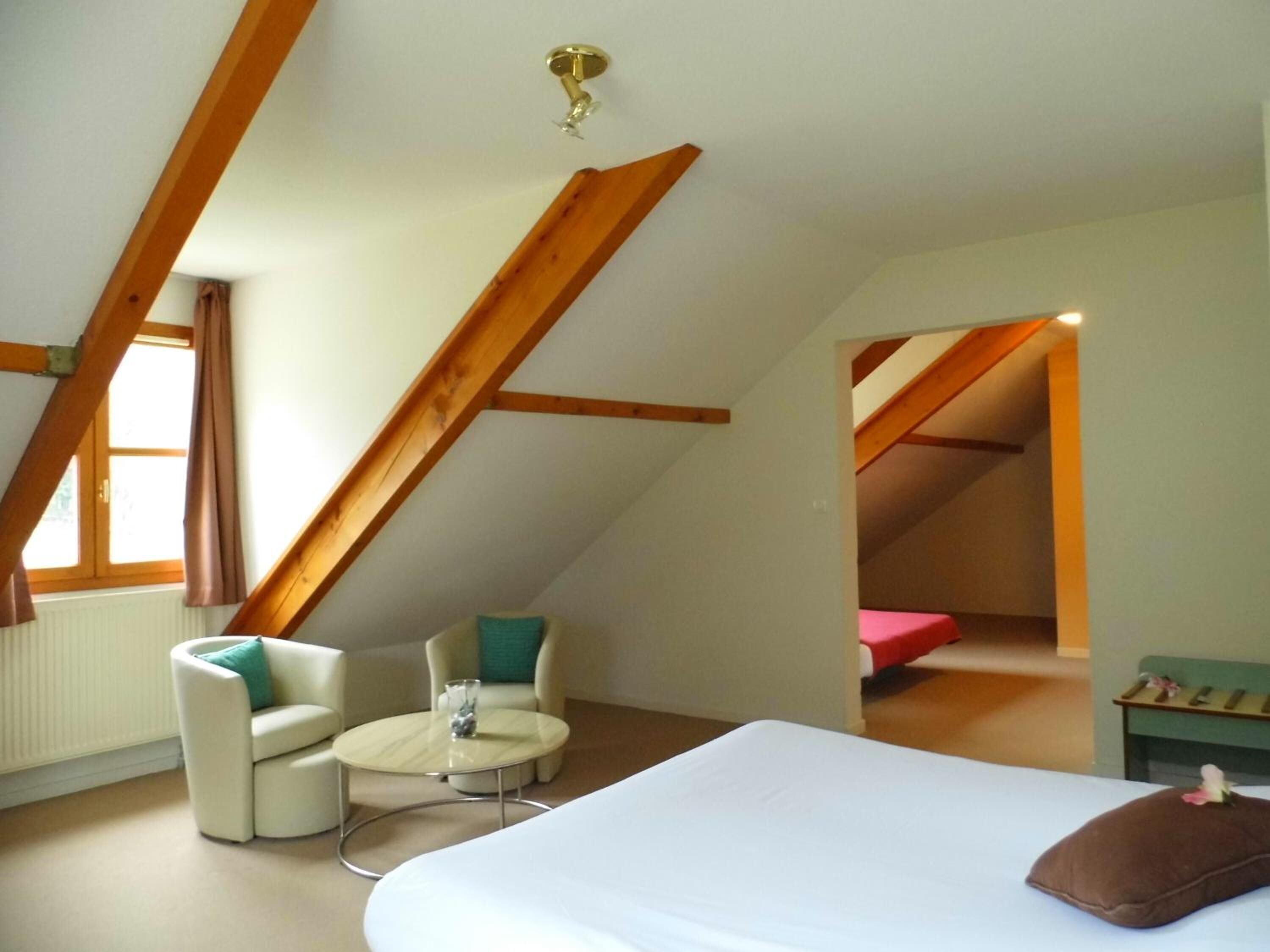 Een slaapkamer met een bed, een klein rond tafeltje, twee stoelen en een raam met gordijnen.