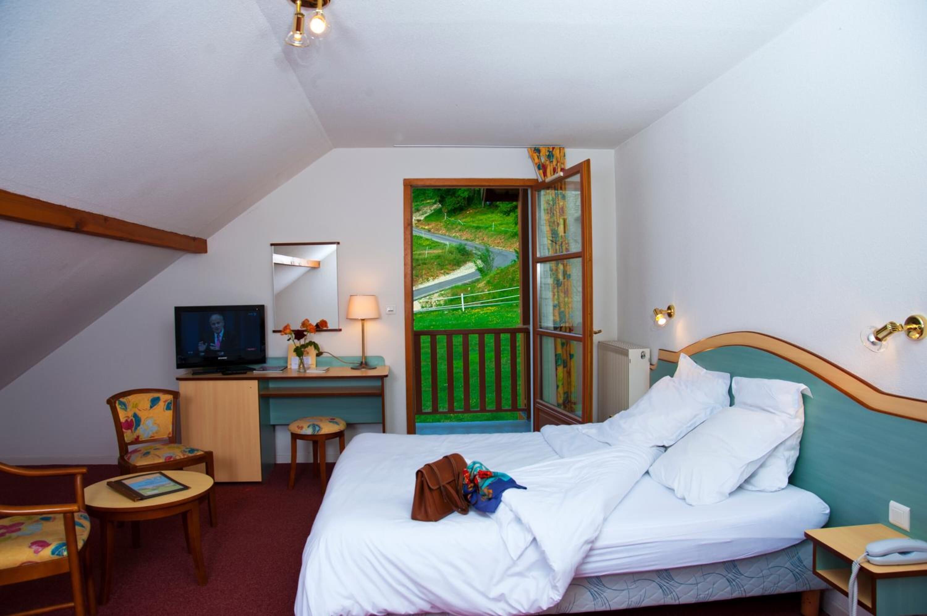 Ein Hotelzimmer mit Bett, Schreibtisch mit Fernseher, Stuhl, kleinem Tisch und Blick auf eine grüne Landschaft durch ein offenes Fenster.