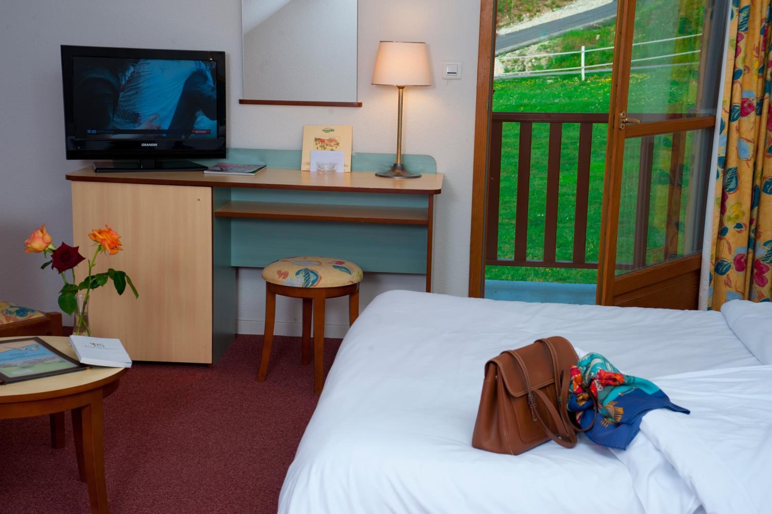 Ett hotellrum med en säng, ett skrivbord med en tv, en lampa, en liten pall och ett fönster med utsikt över grönområden.