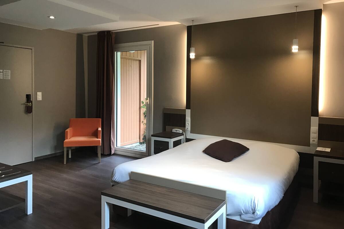 A modern hotel room with a large bed, a desk, a chair, a bedside table, a phone, and a door with curtains.