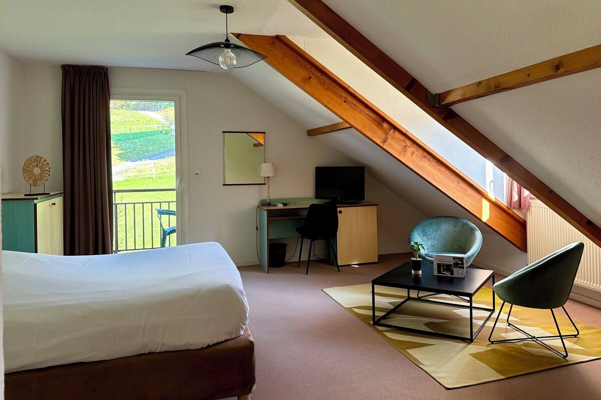 A bedroom with a bed, a desk, a chair, a TV, and a balcony with a view of the outdoors.