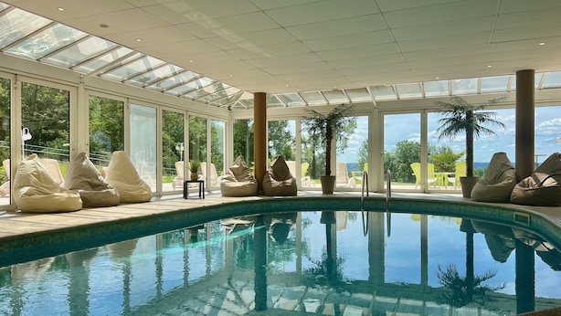 An indoor swimming pool with clear glass walls, beige bean bags, and a view of the outdoors. - Hotel Restaurant Les Bergeronnettes (Champagneux)