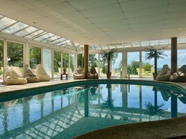 Une piscine intérieure avec des parois en verre transparent, des fauteuils en forme de poire de couleur beige et une vue sur l’extérieur.