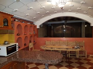 A room with a checkered floor, a round table with a patterned tablecloth, a microwave, and a cabinet with shelves. - Rental Villa Amerchich (Marrakech)