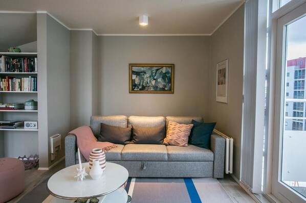 Una sala de estar con un sofá gris, un otoman rosa, una mesa de centro blanca y una estantería llena de libros y objetos decorativos.