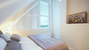 Une chambre avec un plafond en pente, une grande fenêtre, un lit avec des oreillers, une couverture et un tableau accroché au mur.
