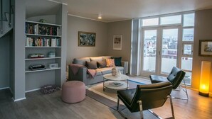 Un salon moderne avec un canapé gris, une table basse, un fauteuil en cuir noir, un pouf rose et une bibliothèque remplie de livres et d’objets décoratifs.