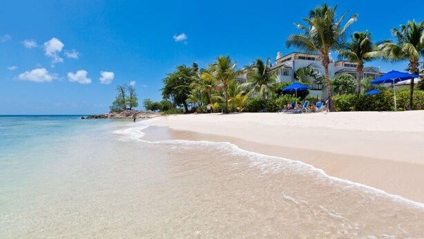 Una playa con agua turquesa cristalina, una orilla de arena y un resort con palmeras y sombrillas azules.