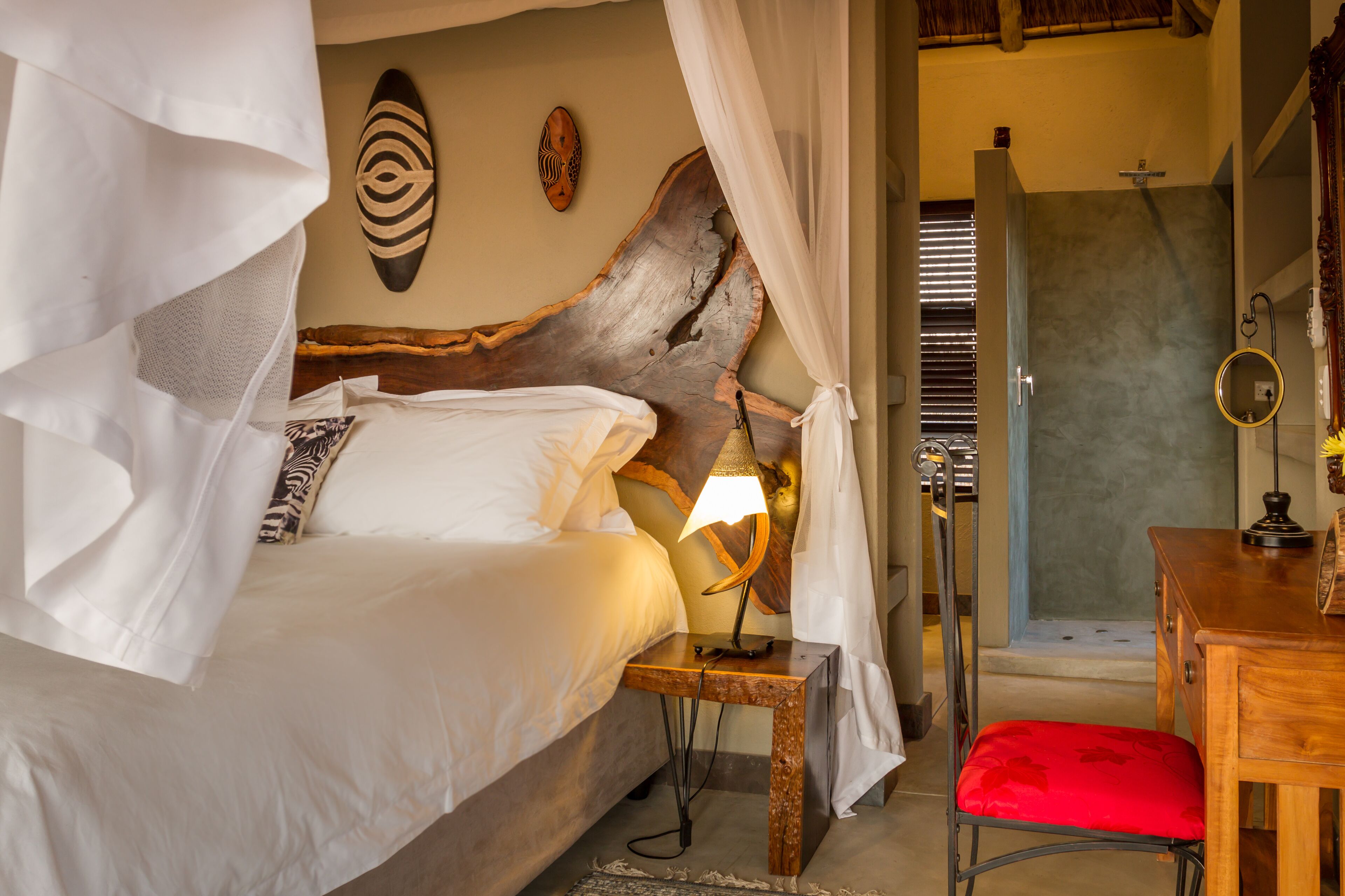 A bedroom with a bed, a nightstand with a lamp, a wooden headboard, a red chair, and a shower area.