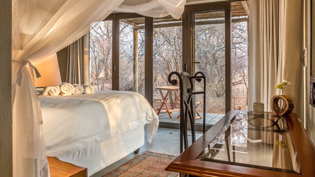 A bedroom with a canopy bed, a glass table, and a view of trees through large windows. - Ukuthula Bush Lodge (Hoedspruit)