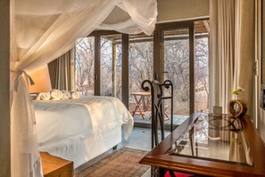 Un dormitorio con cama con dosel, una mesa de vidrio y vista a árboles a través de amplios ventanales.