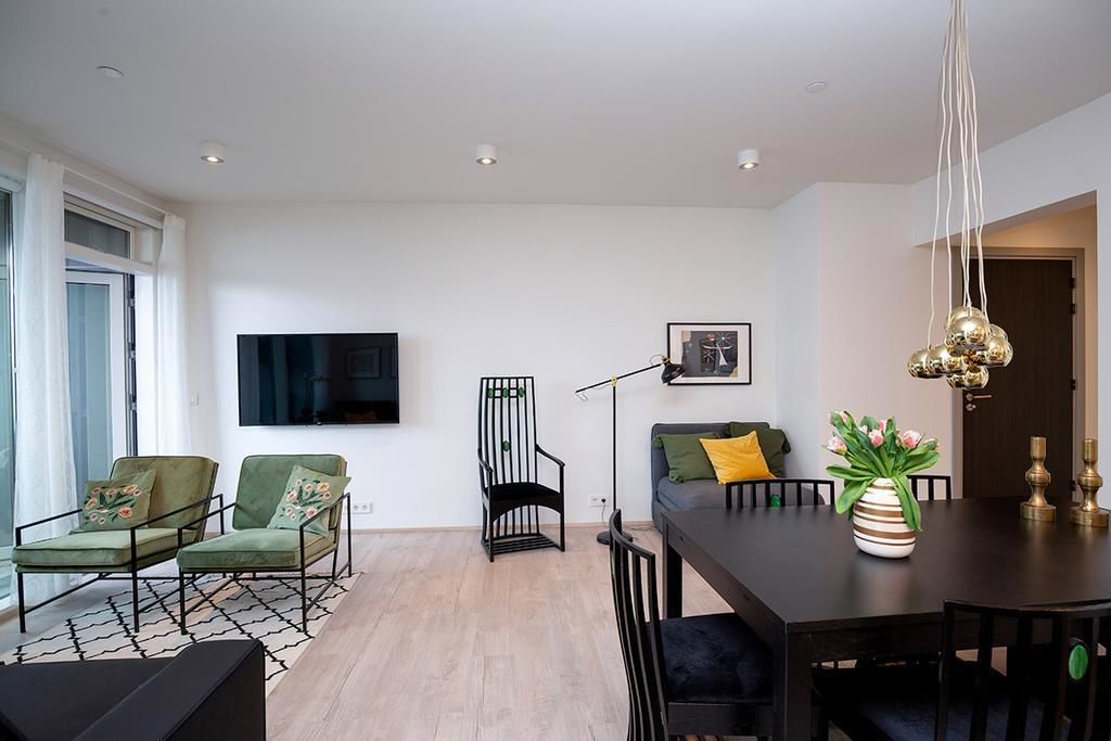 Uma sala de estar moderna com uma mesa de jantar de madeira escura, poltronas verdes, uma cadeira preta e um sofá com almofada amarela. Uma televisão de tela plana está fixada na parede, e há um abajur de chão.