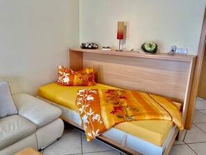 Une petite chambre à coucher moderne, comprenant un lit, un canapé, une table de chevet et une lampe fixée au mur.