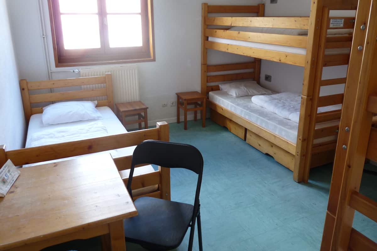 A dormitory room with bunk beds, a wooden desk, a chair, and a window.