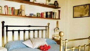 Un dormitori amb un llit de fusta, un edredó blau i una tauleta de nit. Hi ha prestatges sobre el llit amb diversos llibres i objectes decoratius.