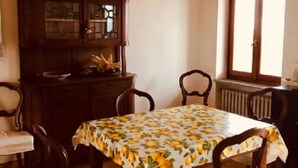 Zona de dinar amb taula coberta per un mantell floral, envoltada d'amb cadira de fusta amb seients amb coixí. Armari de fusta amb portes de vidre contra la paret, i finestra amb vista a l'exterior.